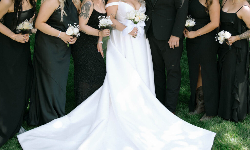 Close-up of the lower half of the bride’s dress train surrounded by bridesmaids in black dresses holding white bouquets at White Barn Weddings in Emmett Idaho
