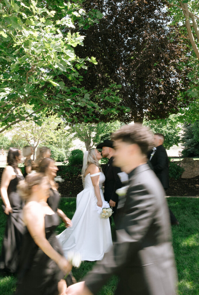 Bride and groom share a kiss under the trees while the wedding party in motion blurs around them at White Barn Weddings in Emmett Idaho