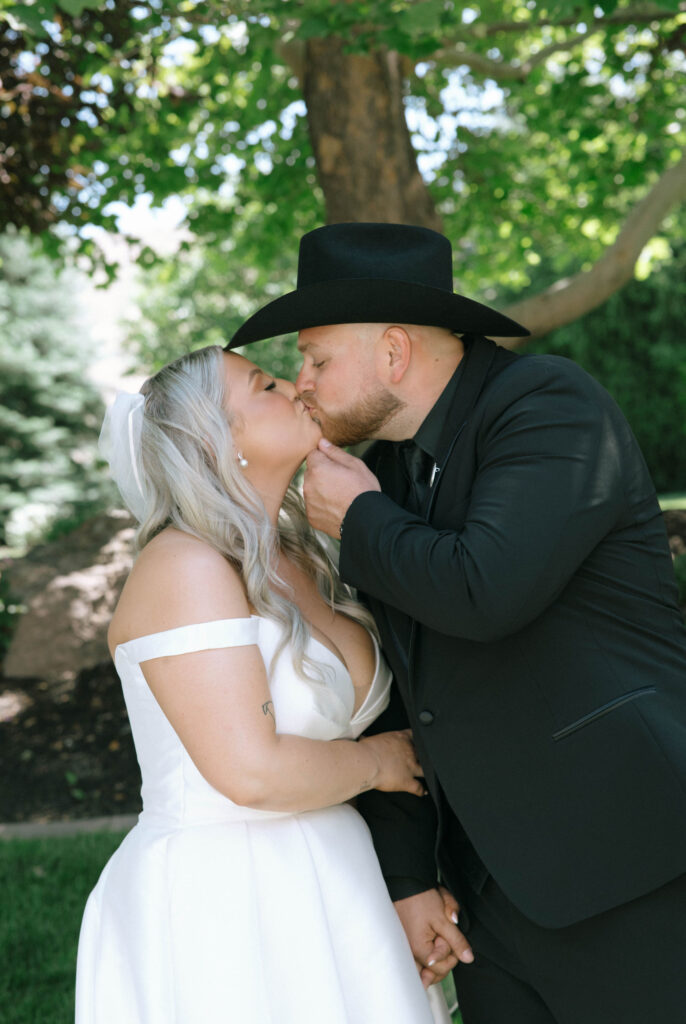 Bride and groom kissing in a shaded garden area, surrounded by tall trees and lush grass at White Barn Weddings in Emmett, Idaho
