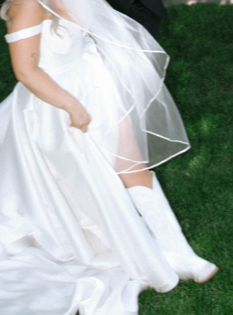 Bride lifting her bridal dress to reveal white cowboy boots, veil flowing in the breeze at White Barn Weddings in Emmett, Idaho