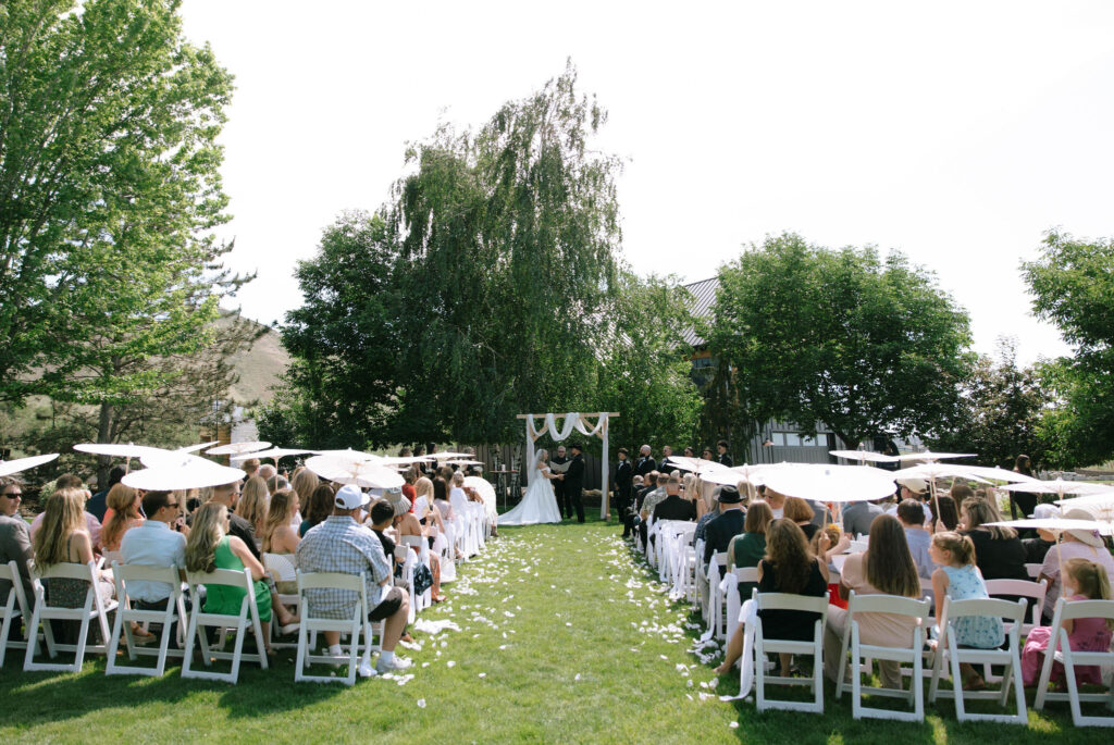 Wide view of White Barn Weddings in Emmett Idaho outdoor ceremony with guests seated under parasols and the couple at the altar.