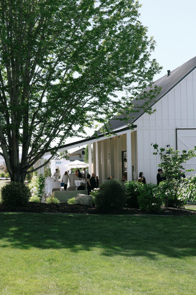 Exterior of White Barn Weddings in Emmett, Idaho on a sunny June day, with guests mingling on the shaded veranda surrounded by lush green landscaping, captured by Krysta Timmons Photography