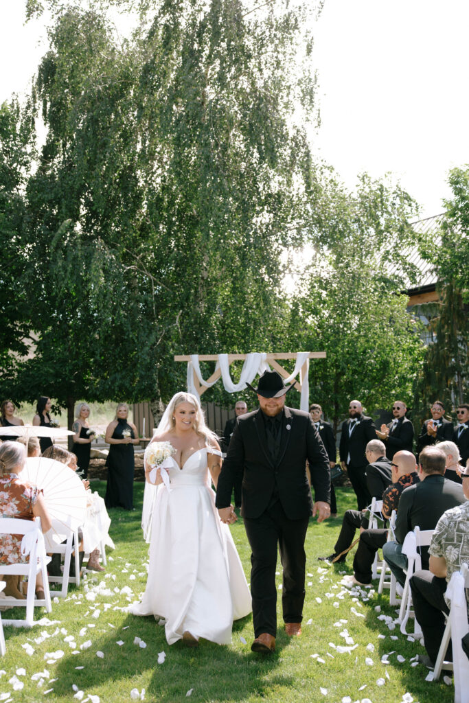 Newly married couple walking down the aisle smiling and holding hands, with bridal party clapping in the background at White Barn Weddings in Emmett Idaho