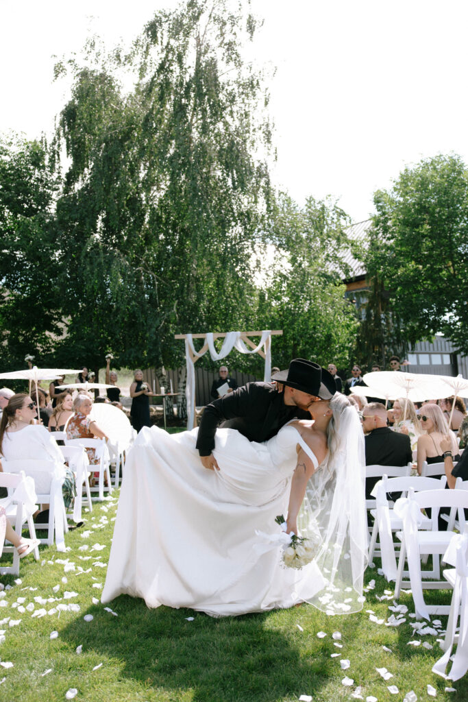 Groom dipping the bride for a romantic kiss in the aisle, surrounded by guests and scattered white rose petals at White Barn Weddings in Emmett Idaho