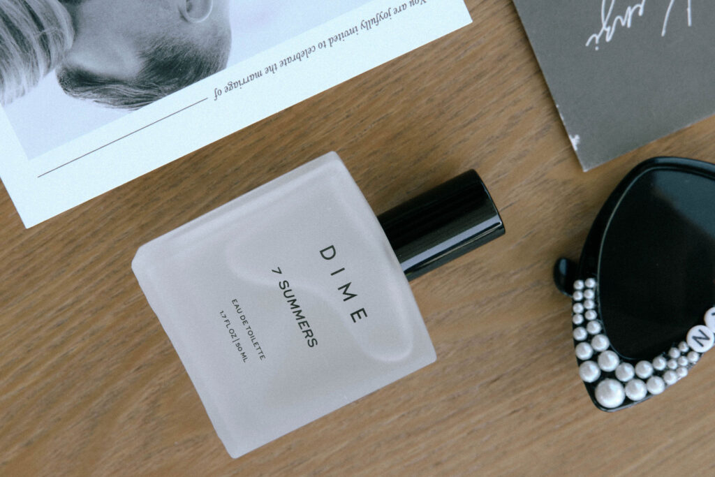 Detail shot of groom’s cologne, DIME 7 Summers, placed beside wedding invitation and black sunglasses with pearl embellishments
