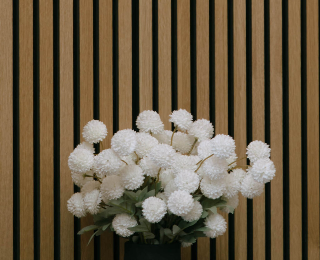 Modern floral arrangement of white pom-pom flowers in a black vase, displayed against vertical wooden slat wall at White Barn Weddings venue in Emmett, Idaho