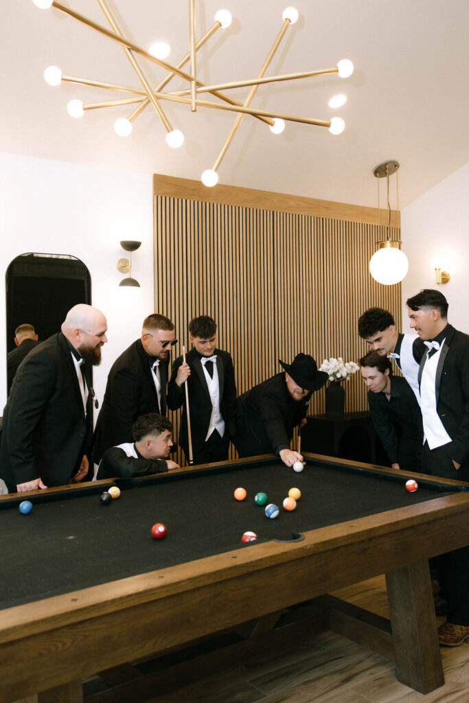 Groomsmen in black tuxedos playing pool in the modern groom’s suite at White Barn Weddings in Emmett, Idaho, captured by Krysta Timmons Photography