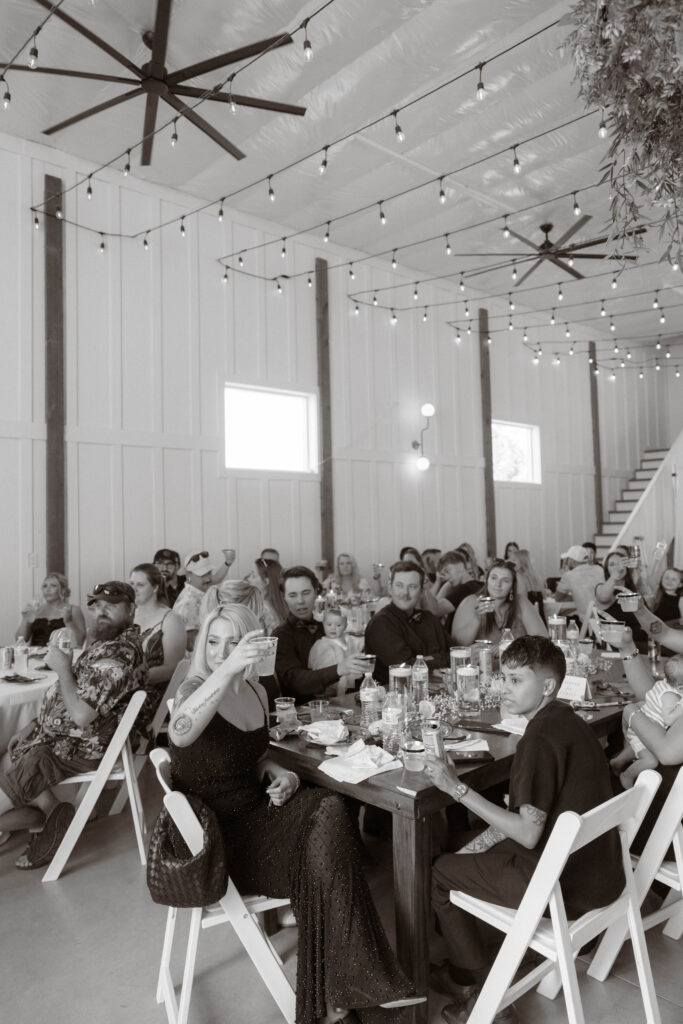 Black-and-white candid of wedding guests raising glasses for a toast inside White Barn Weddings in Emmett Idaho