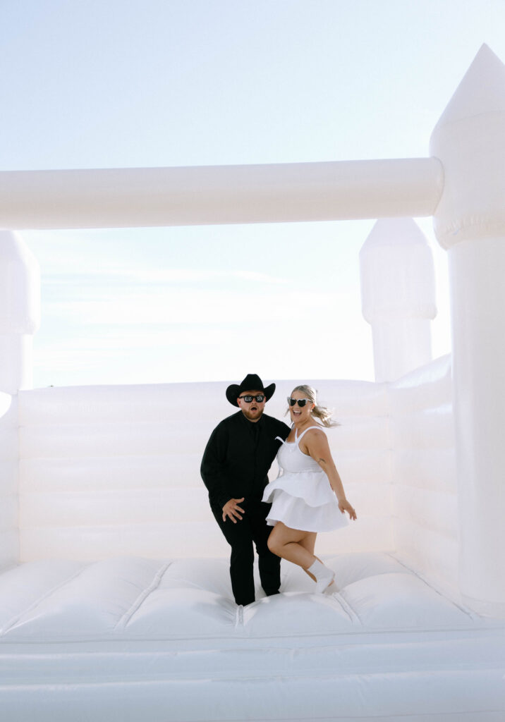 Bride and groom jumping on white inflatable bounce house at wedding reception, groom in black cowboy hat and sunglasses, bride in short white dress with heart-shaped sunglasses at White Barn Weddings in Emmett, Idaho