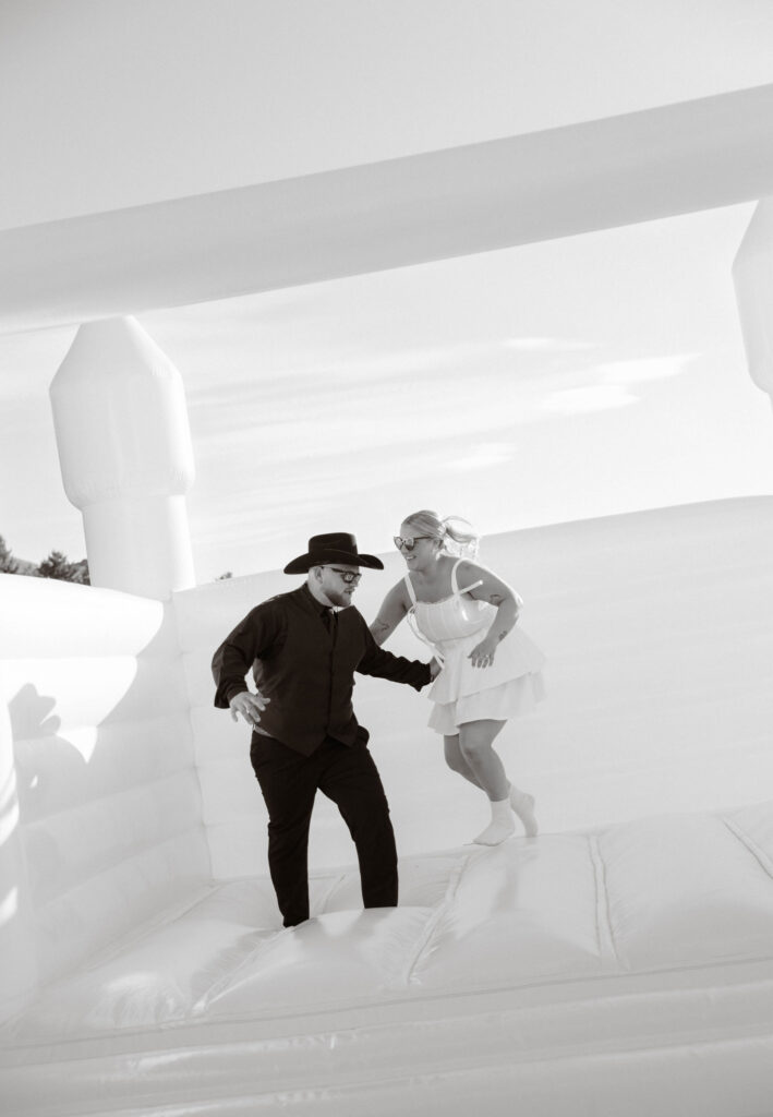 Black-and-white photo of bride and groom laughing and bouncing together on wedding inflatable, playful moment at outdoor reception at White Barn Weddings in Emmett, Idaho