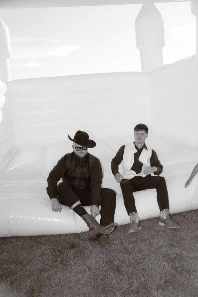 Black-and-white photo of groom in cowboy hat and sunglasses sitting on inflatable with groomsman in white vest, relaxed candid wedding reception moment at White Barn Weddings in Emmett, Idaho