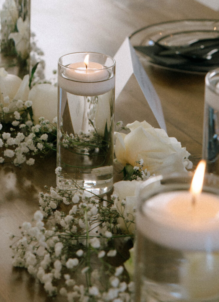 Close-up of floating candle centerpieces in tall glass vases, surrounded by white roses and baby’s breath on wooden reception tables at The White Barn